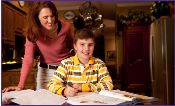 Boy smiles at the camera while doing schoolwork, with his mom standing behind him